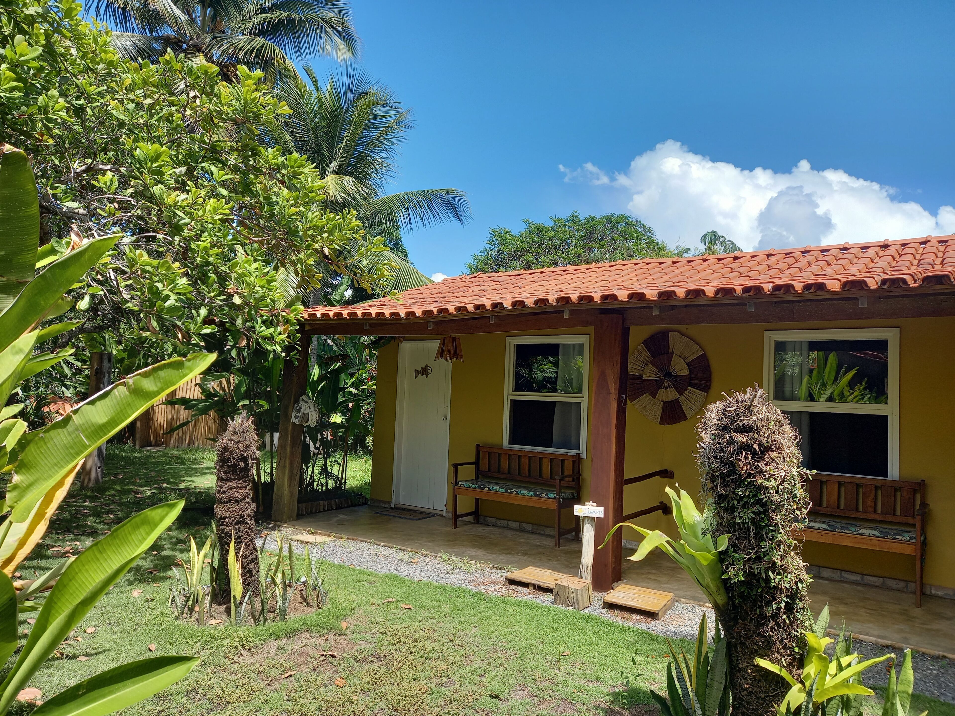A small yellow building with a tiled roof, two windows, and a wooden bench outside.