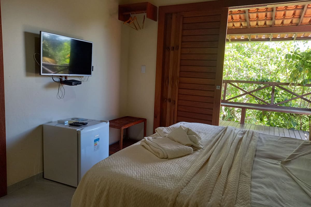 A bedroom with a bed, a television, a small refrigerator, and a window with a view of greenery.