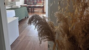 Ein Zimmer mit Holzboden, einer Couch, einem Bett mit grün kariertem Überwurf, einem Spiegel und einem Fenster mit Vorhängen.