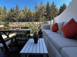 Un patio al aire libre con una mesa blanca, bancos de madera, un sofá blanco con cojines rojos y un muro de piedra de fondo.