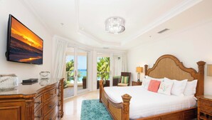 A spacious bedroom with a large bed, a dresser, a TV mounted on the wall, and a view of the ocean through the sliding doors.