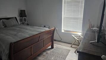 A bedroom with a wooden bed, a desk with a computer, and a window with blinds.
