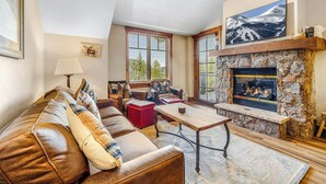 A cozy living room with a stone fireplace, leather sofa, and a large TV mounted above the fireplace. - Mountain Thunder Bluebird Condo: Ski In, Top-Floor w/ Views, Resort Amenities (Breckenridge)