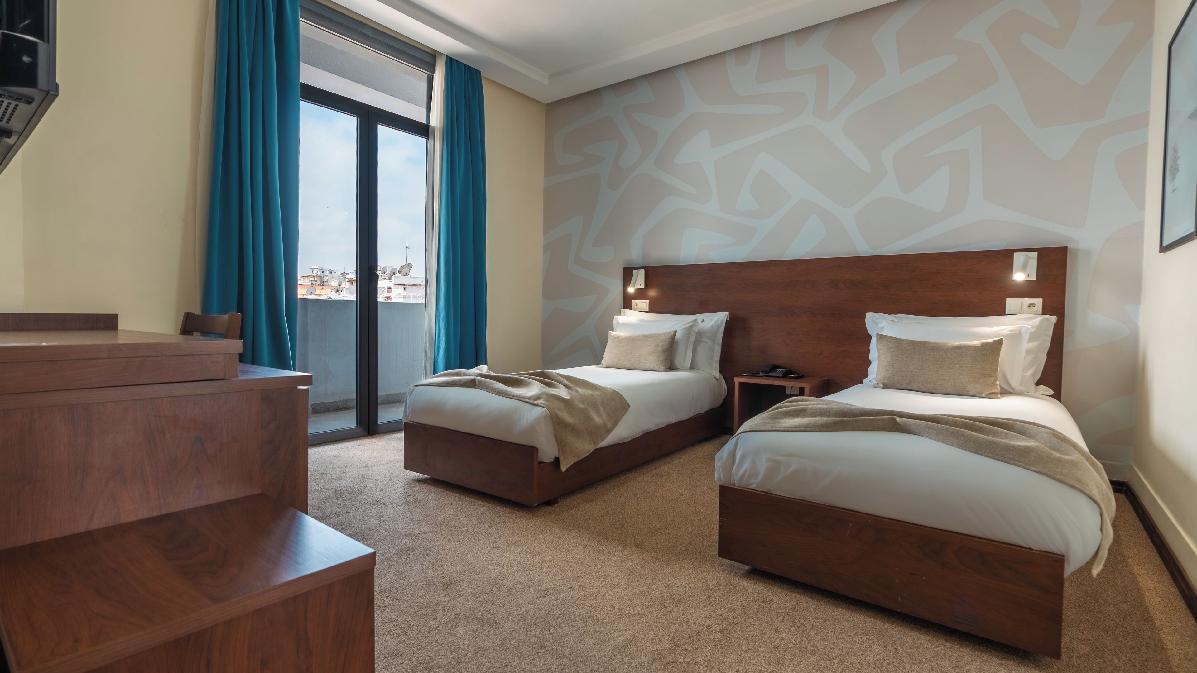 A hotel room with two beds, a desk, and a view of the city through a large window.