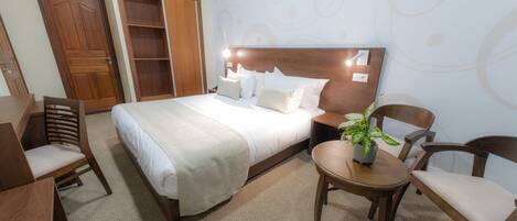 A hotel room with a large bed, wooden headboard, a bedside table with a lamp, a small round table with a chair, and a potted plant.