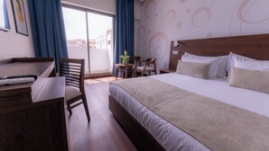 Ein Hotelzimmer mit einem großen Bett, einem Schreibtisch und einem Stuhl. Ein Fenster mit Vorhängen, eine Wand mit abstrakten Mustern und Blick auf Gebäude.