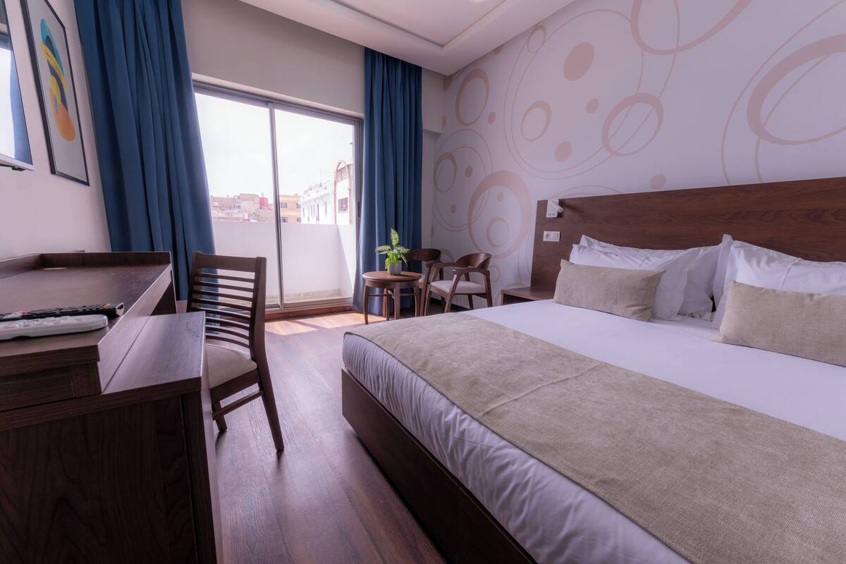 Une chambre d’hôtel avec un grand lit, un bureau et une chaise. Il y a une fenêtre avec des rideaux, un mur orné de motifs abstraits et une vue sur les bâtiments à l’extérieur.