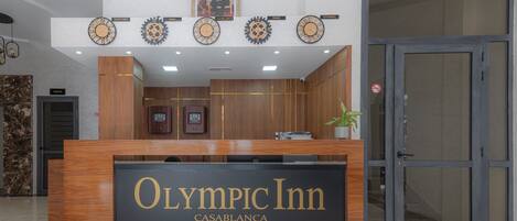 Un mostrador de recepción con un cartel que dice "Olympic Inn Casablanca" y relojes que muestran la hora de Nueva York, París e Estambul.