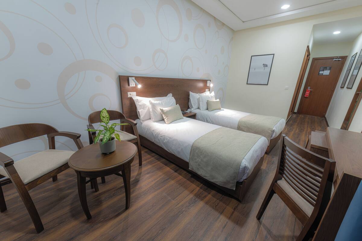 Une chambre d’hôtel avec deux lits, une petite table avec une plante, une chaise et un bureau.