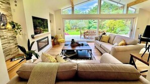 A spacious living room with a large sofa, a coffee table, and a television. There are large windows providing a view of the outdoors. - Spacious Stylish Forest Escape | Kids Outdoor Play (Saint Leonards)