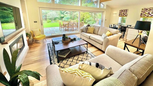A spacious living room with a large window, a fireplace, and a dining area with outdoor seating. - Spacious Stylish Forest Escape | Kids Outdoor Play (Saint Leonards)