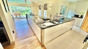 A modern kitchen with a central island, a dining area with a table and chairs, and a view of the outdoors. - Spacious Stylish Forest Escape | Kids Outdoor Play (Saint Leonards)