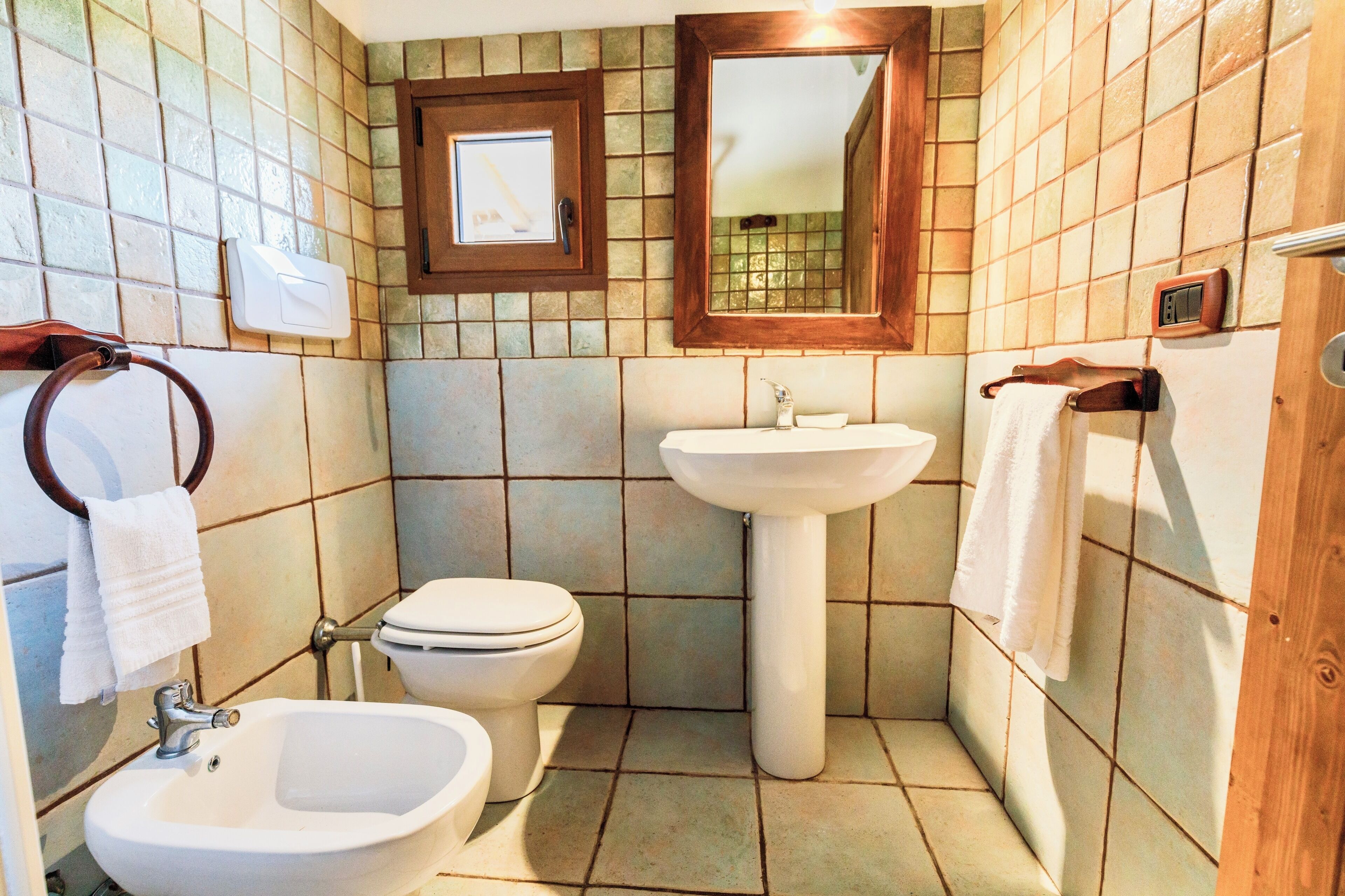 A bathroom with a toilet, bidet, and sink, all with white fixtures, and a wooden-framed mirror above the sink.