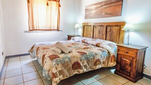 Un dormitorio con cama, cabecera de madera, dos mesitas de noche con lámparas, una ventana con cortinas y un cuadro en la pared.