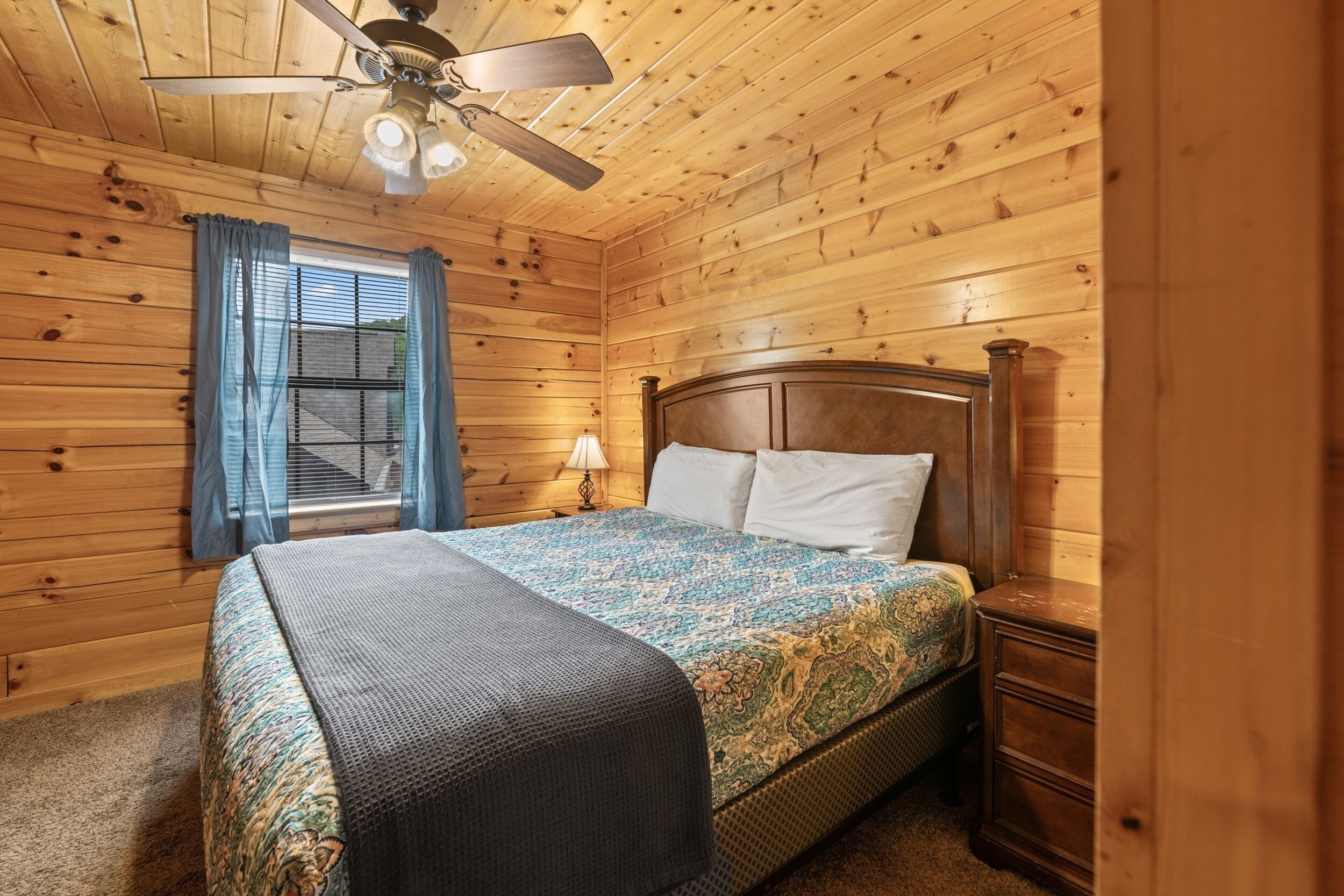 A wooden-paneled bedroom with a bed, a ceiling fan, and a window with curtains.