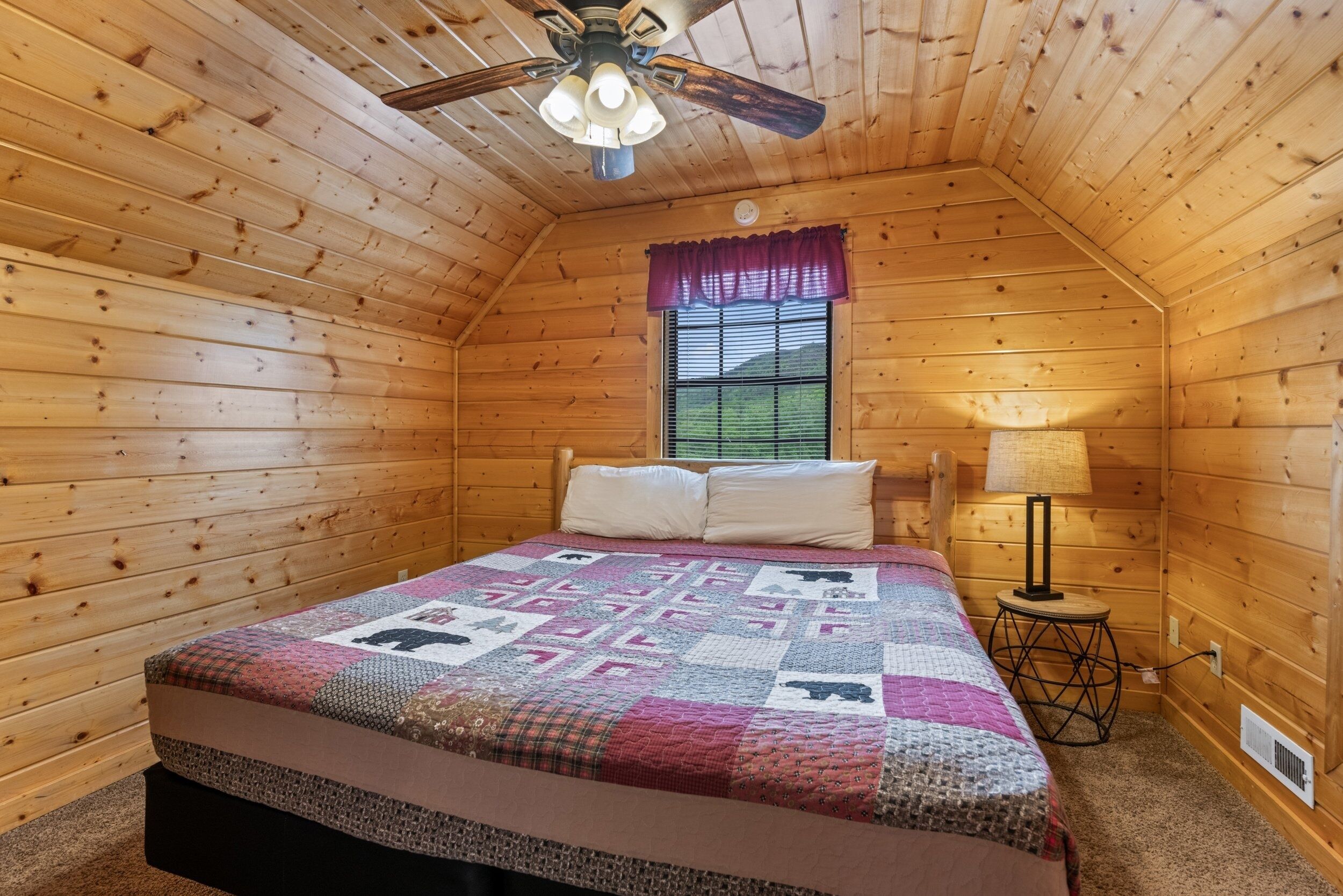 A cozy wooden cabin bedroom with a bed, a window with blinds, a ceiling fan, and a small table with a lamp.