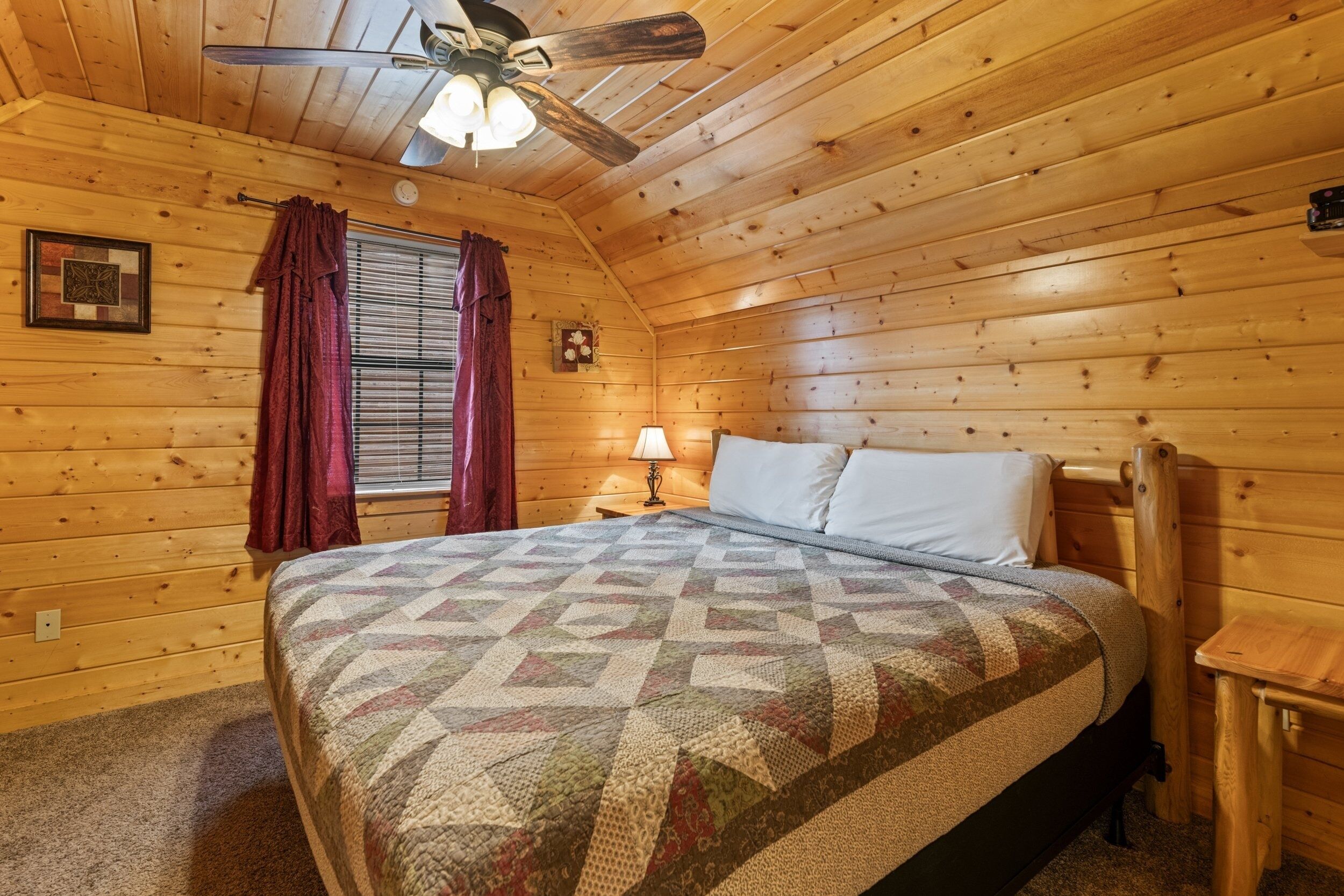 Habitació acollidora d'una cabana de fusta amb llit, ventilador de sostre, finestra amb cortines i una taula petita.