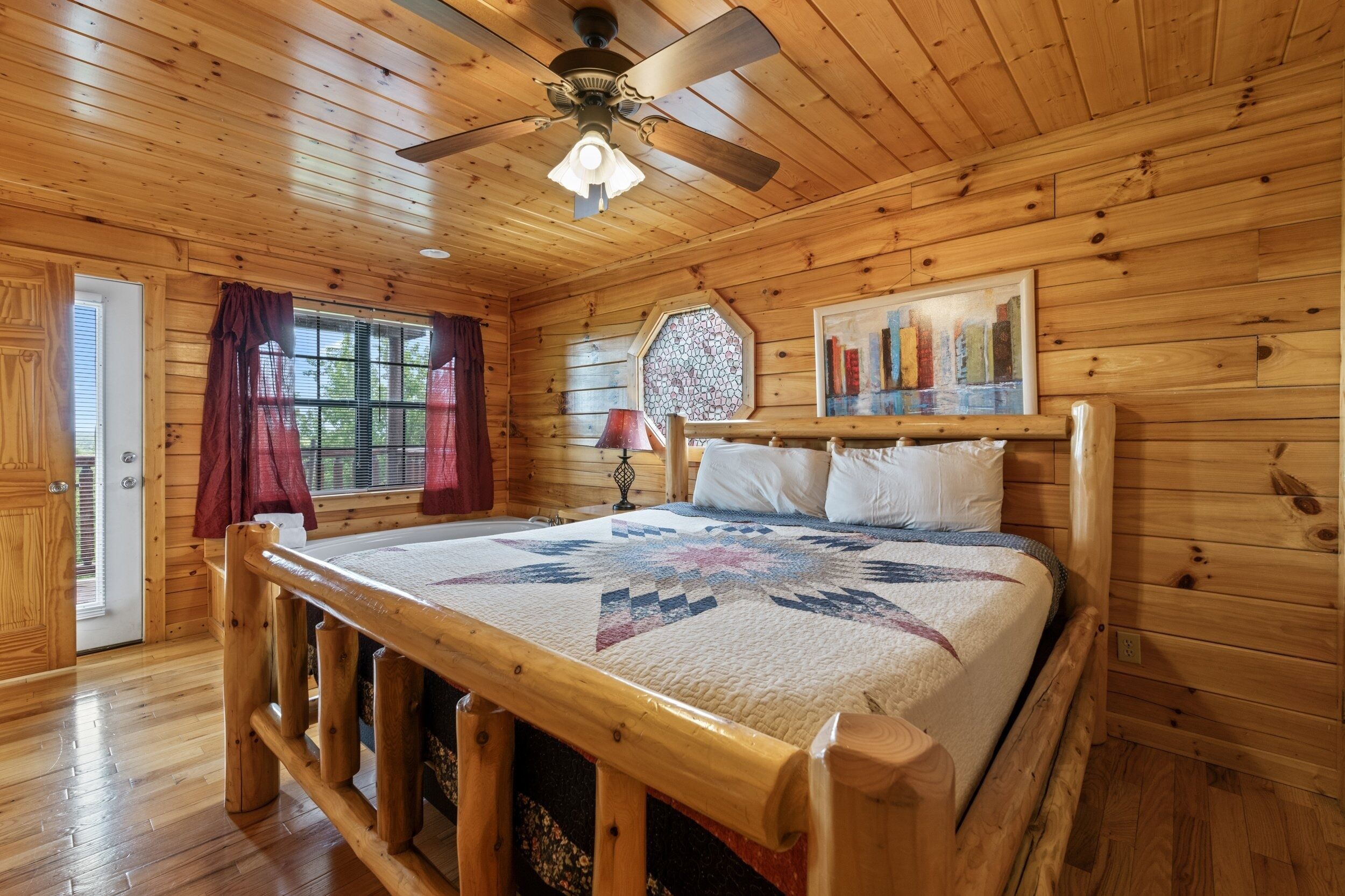 A wooden cabin bedroom with a bed, a ceiling fan, a window with curtains, and a painting on the wall.