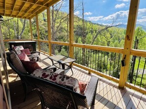 Un espace extérieur aménagé, abrité et doté de meubles en osier et d’une table en verre, offrant une vue sur un paysage boisé.