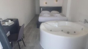 A modern bathroom with a large bathtub, a small table with a chair, and a bed with pillows. - Verykokkos on the Beach (Naxos)