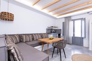 A modern living room with a sofa, dining table, and chairs. There is a kitchen area with a sink and a window with curtains.