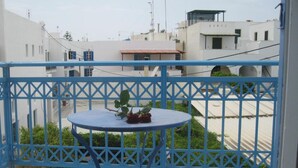 A balcony with a blue metal railing, a small round table with a plant, and a view of a residential area with white buildings and a red-roofed structure. - Verykokkos Pension 2 (Naxos)