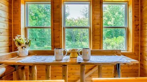 A wooden cabin interior with a table set for two, two mugs, a vase with flowers, and a window with a forest view.