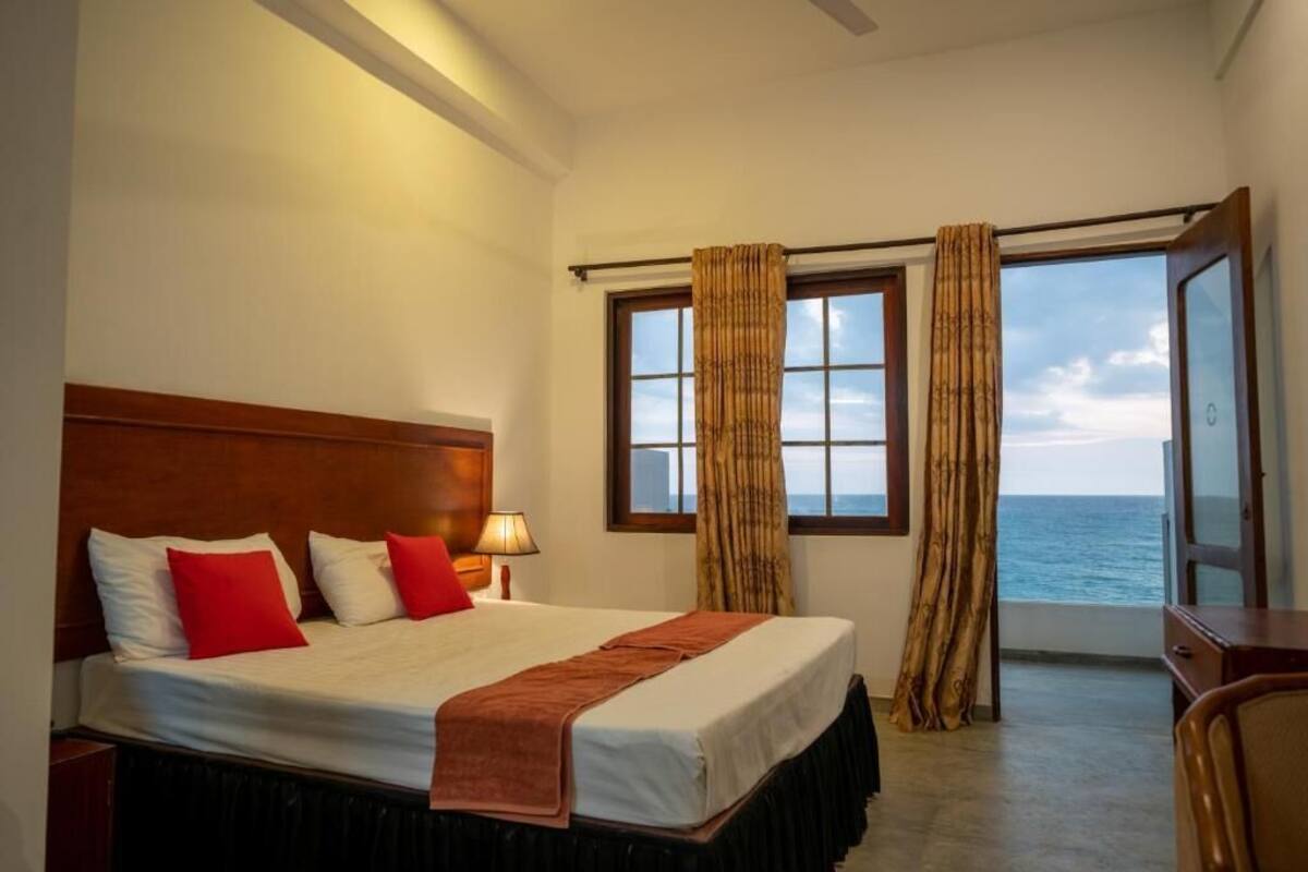 Une chambre d’hôtel avec un grand lit, une tête de lit en bois, une table de chevet, une lampe et une vue sur la mer par une fenêtre avec des rideaux.