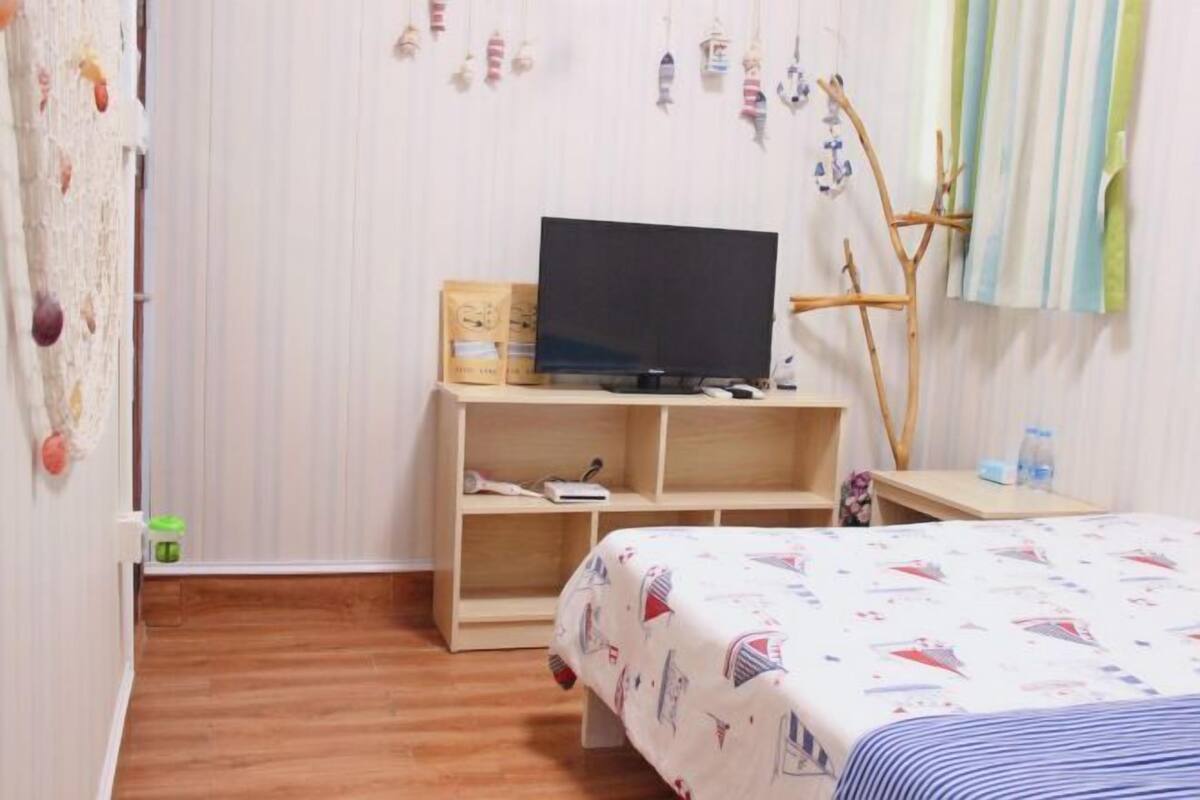 A bedroom with a bed, a TV on a wooden stand, a shelf with items, and a window with curtains.