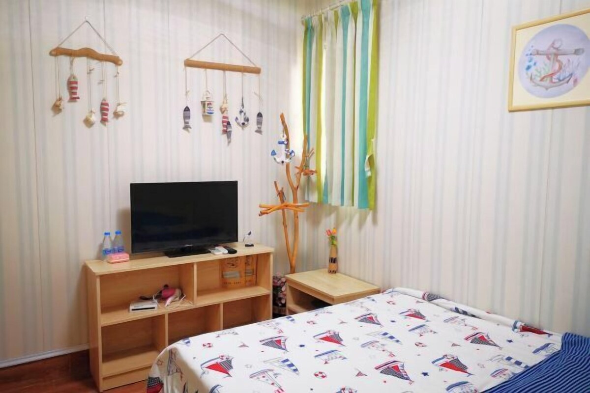 A bedroom with a bed, a TV on a wooden stand, a small table, and a wall with curtains and hanging decorations.