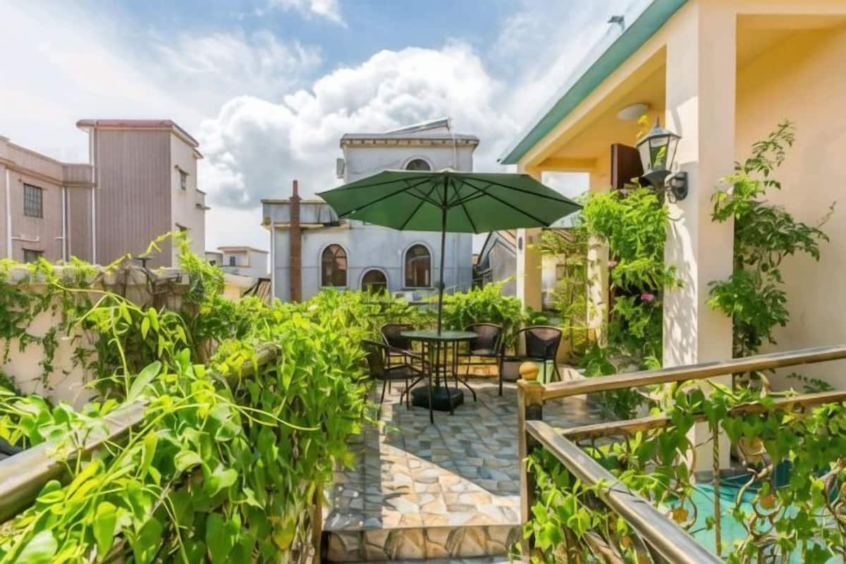 A courtyard with a stone floor, a table with chairs, a green umbrella, and a building with arched windows in the background.