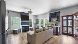 A spacious living room with a beige sofa, a fireplace, and a kitchen area with a wine rack. - Sip by the Pool - One bedroom, Pool, Beach Access (Pawleys Island)