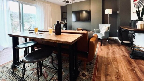 Una sala de estar moderna con una mesa de comedor de madera, un sofá, un televisor montado en la pared y un amplio ventanal con cortinas.
