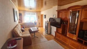 A living room with a sofa, a coffee table, a TV, wooden cabinets, and a kitchen area with a refrigerator and microwave. - Mitten im Thüringer Wald, Ferienhaus für die Ganze Familie (Steinbach-Hallenberg)