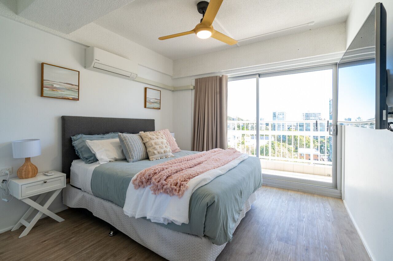 A modern bedroom with a bed, a nightstand, a ceiling fan, and a large window offering a city view.