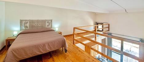 A bedroom with a bed, a nightstand, a wooden desk, and a balcony with a railing.