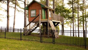 A wooden treehouse with a green door and a staircase leading up to it, surrounded by a black metal fence and tall trees. - One Acre Lakefront Home, Indian Hills Peninsula.  Pet friendly ! (Livingston)