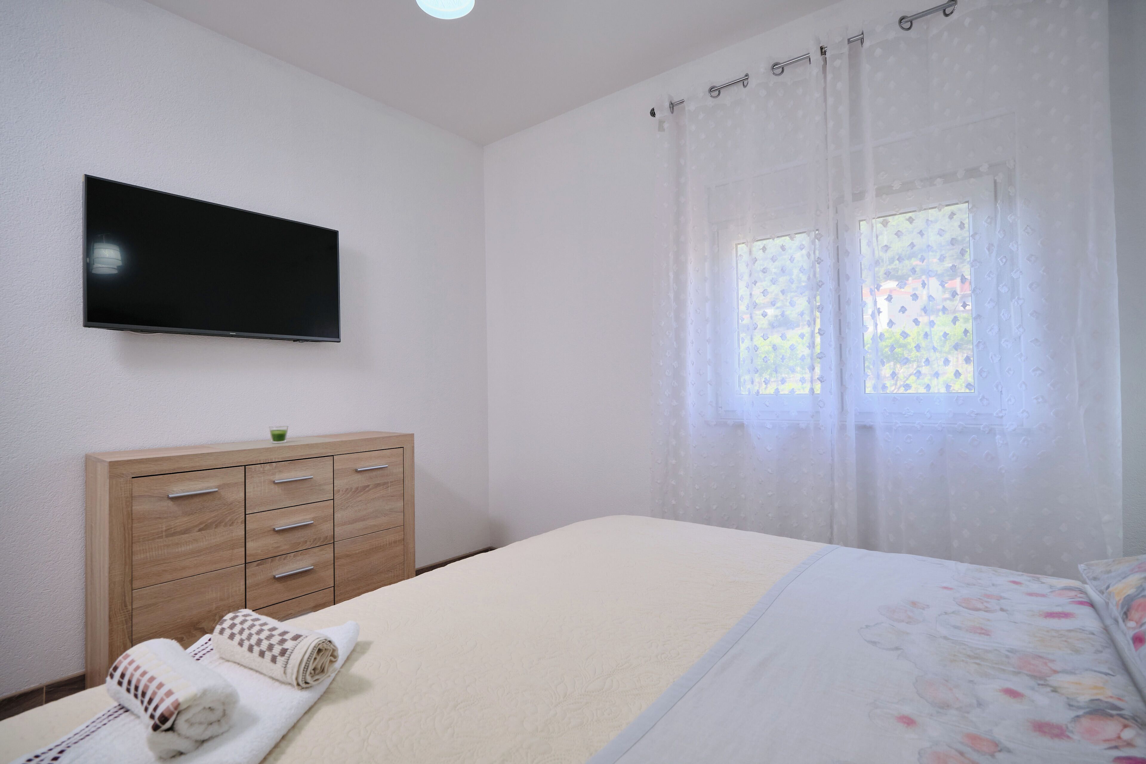 Un dormitorio con cama, televisor montado en la pared, cómoda de madera y ventana con cortinas ligeras.