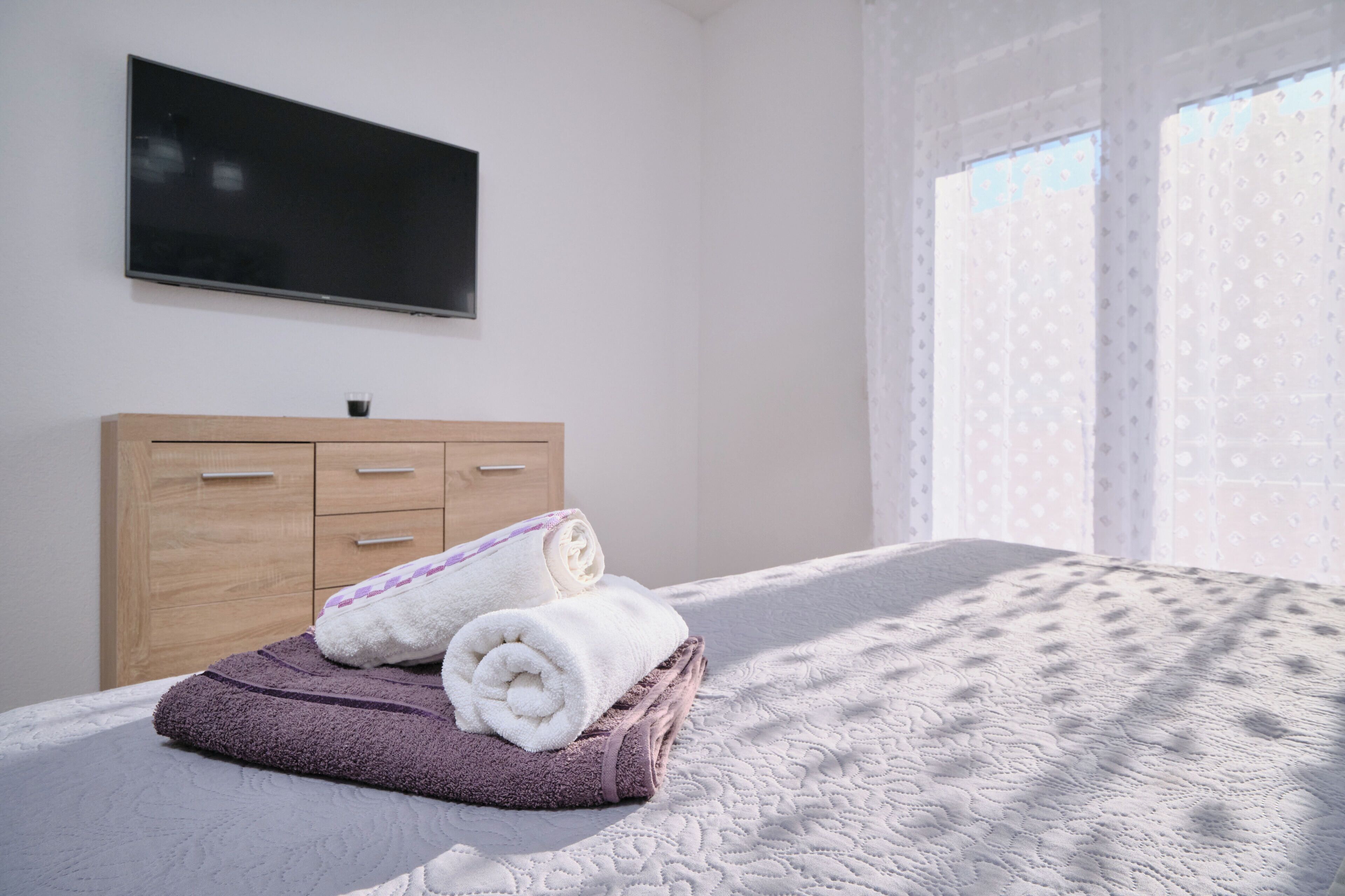 Ein Schlafzimmer mit einem Bett, einem an der Wand befestigten Fernseher, einer Holzkommode und einem Fenster mit gemusterten Vorhängen.