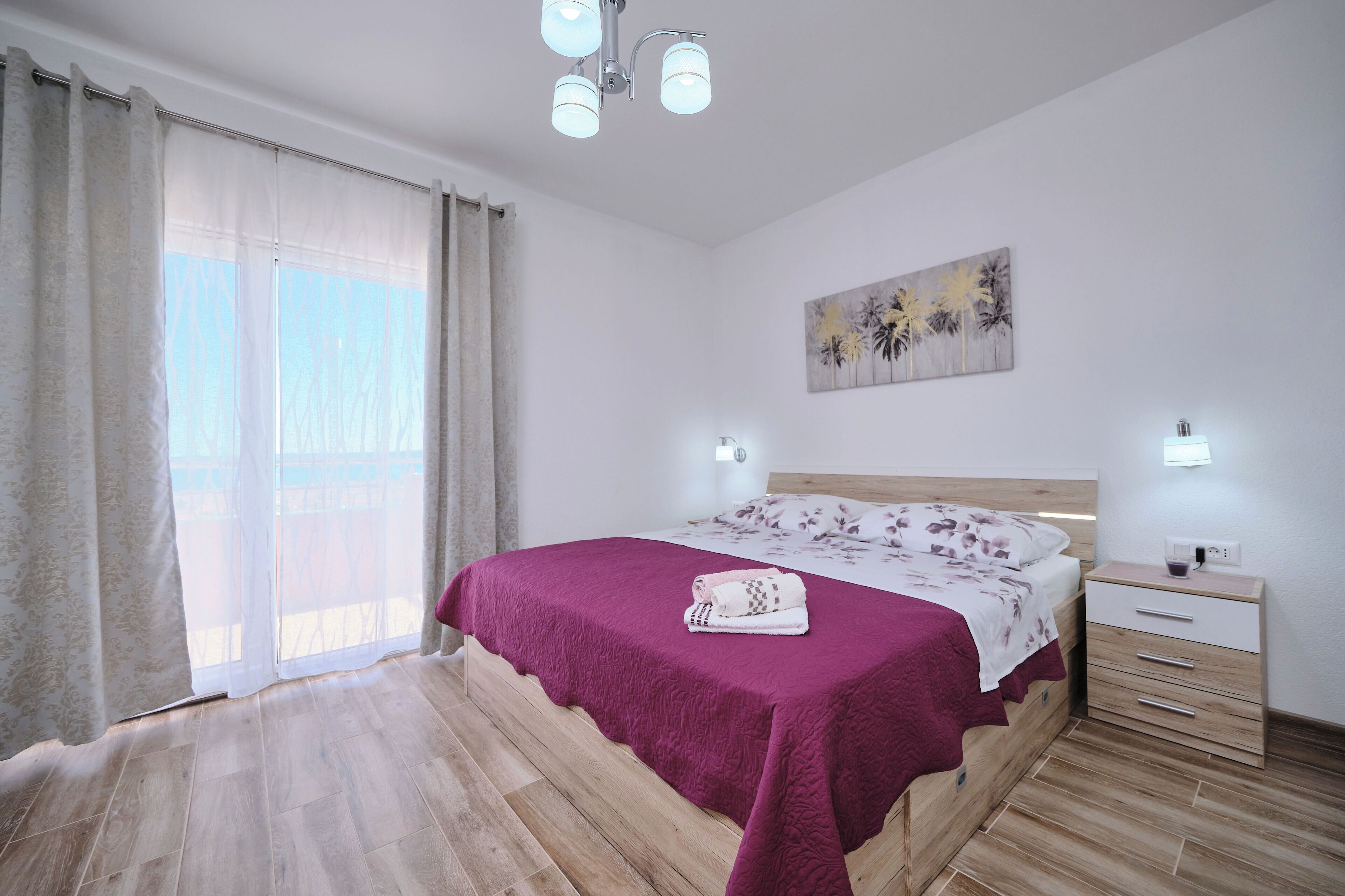 A bedroom with a wooden floor, a bed with a purple bedspread, a nightstand with a lamp, and a painting on the wall.