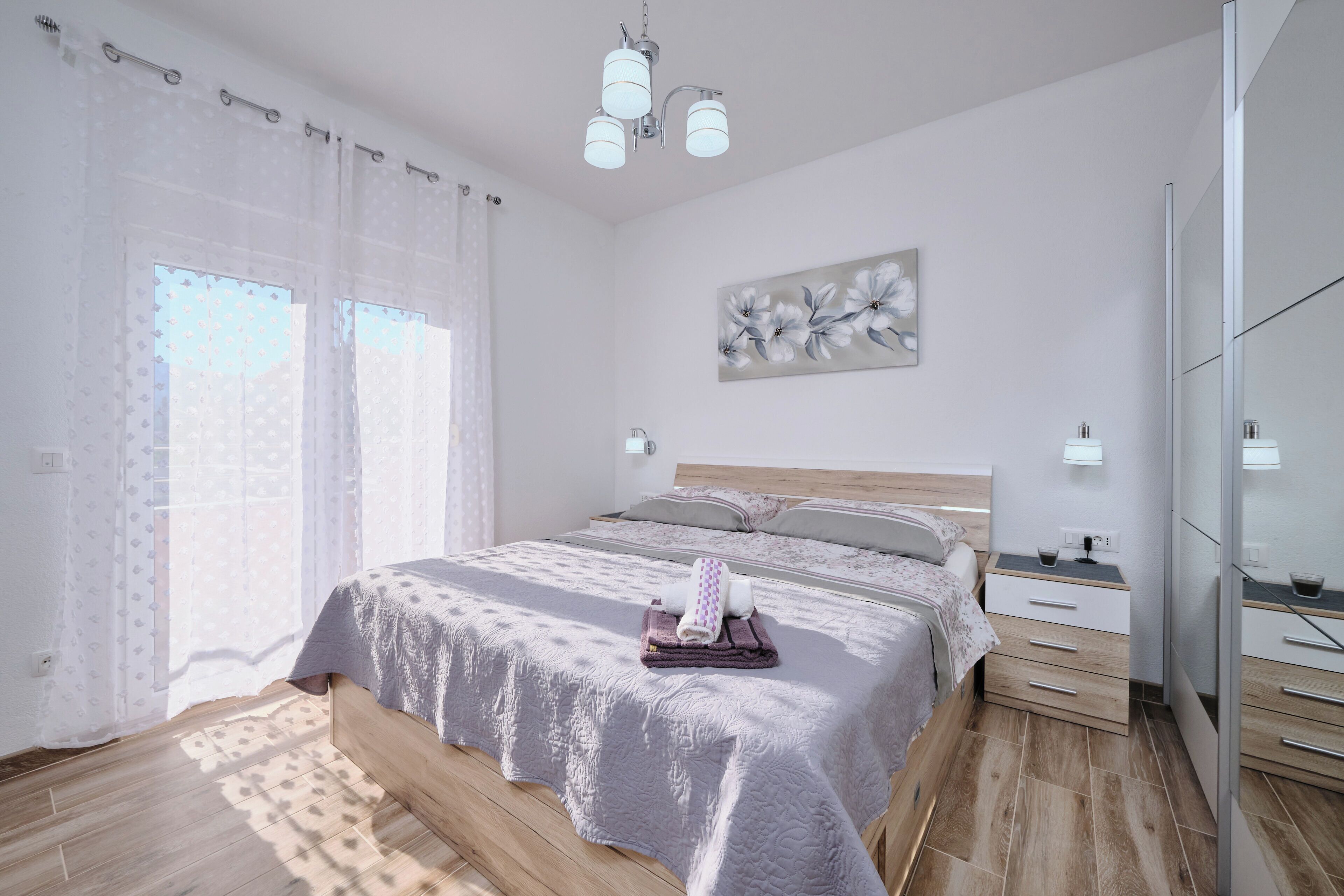 A modern bedroom with a large bed, bedside tables, a mirror, and a window with sheer curtains.