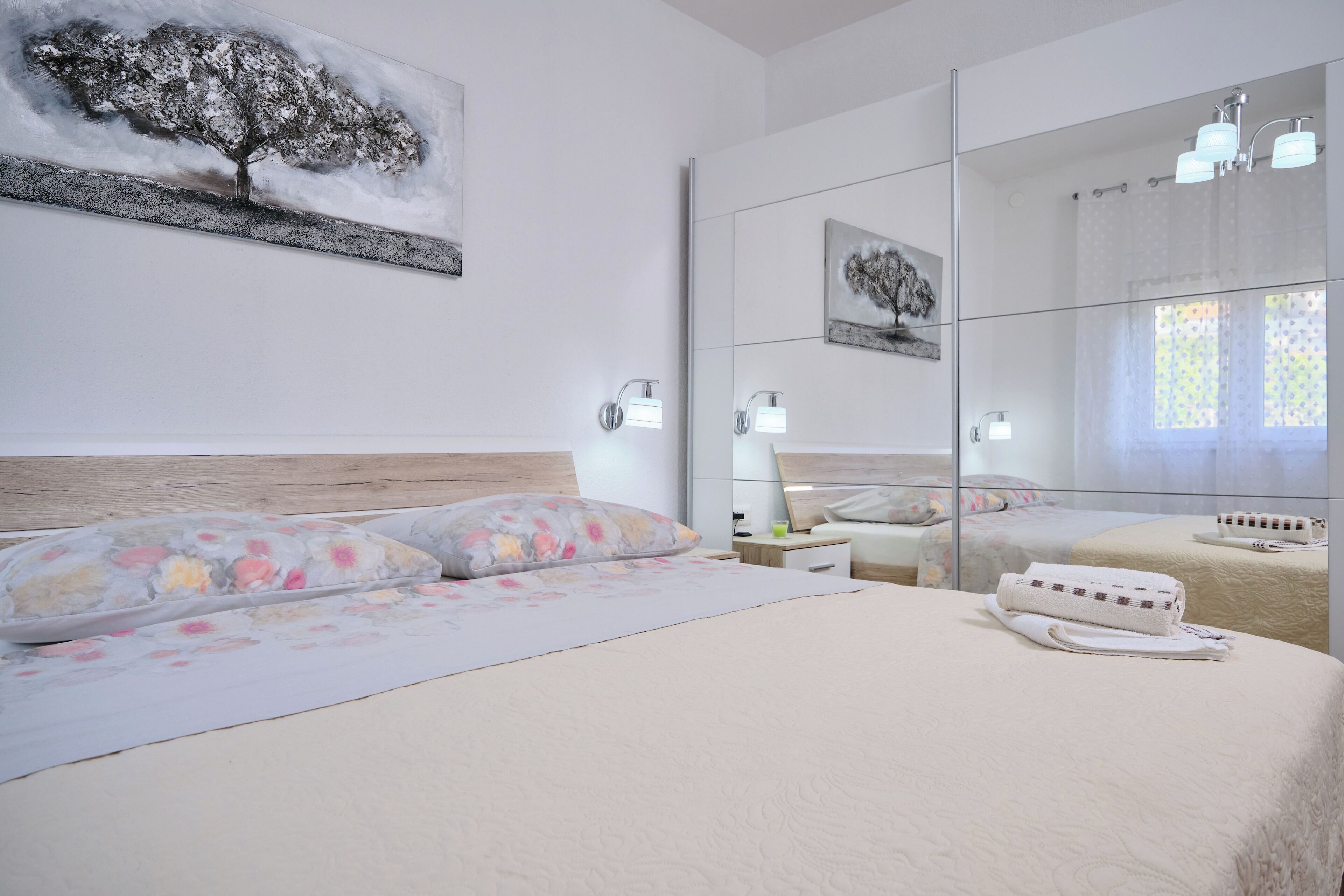 Un dormitorio con una cama, una mesita de noche, un espejo y un cuadro de un árbol.