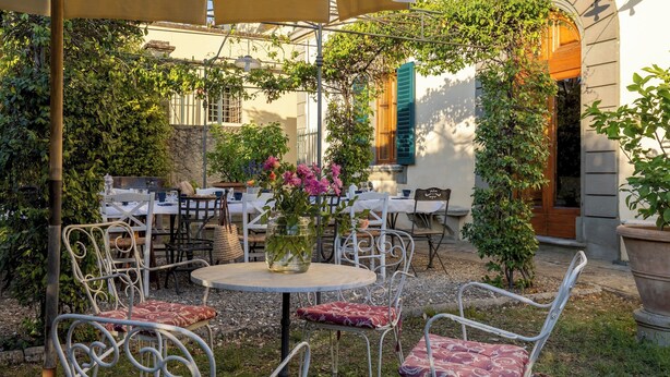 An outdoor café with round tables and metal chairs, surrounded by greenery and a stone wall.