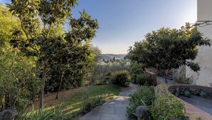 A garden pathway leading to a stone wall with vases, surrounded by lush greenery and trees.