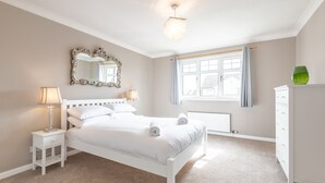 A bedroom with a white bed, a nightstand with a lamp, a dresser, a window with curtains, and a mirror on the wall. - Smugglers' Gait | Anstruther (Anstruther)