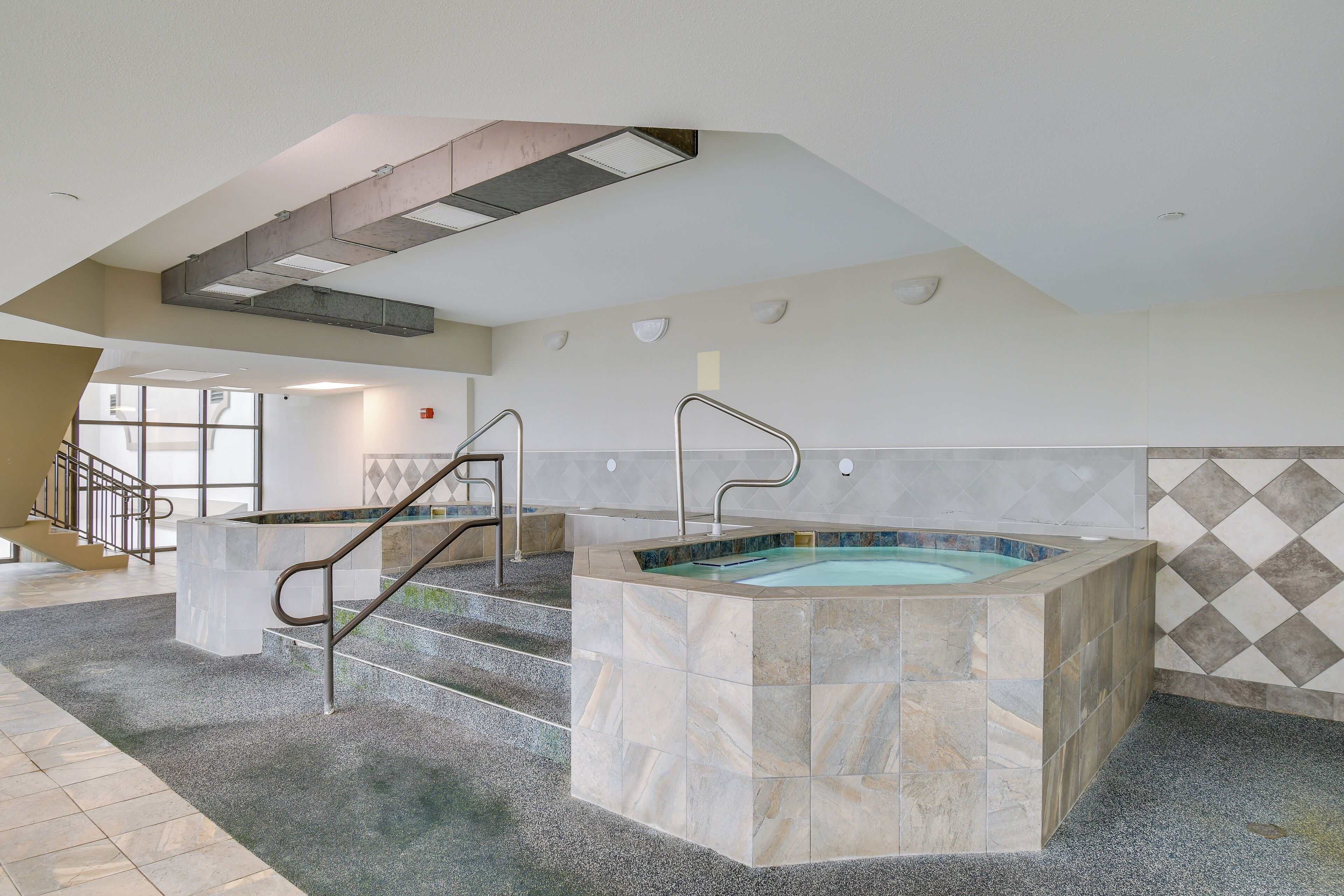 A modern indoor pool area with a central hot tub, a staircase, and a view of the outside.