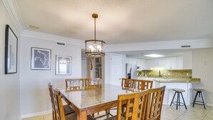 A dining area with a glass-top table and wooden chairs, a kitchen with white cabinets, and a hanging light fixture. - Waterfront Pool: Orange Beach Condo w/ Views! (Orange Beach)
