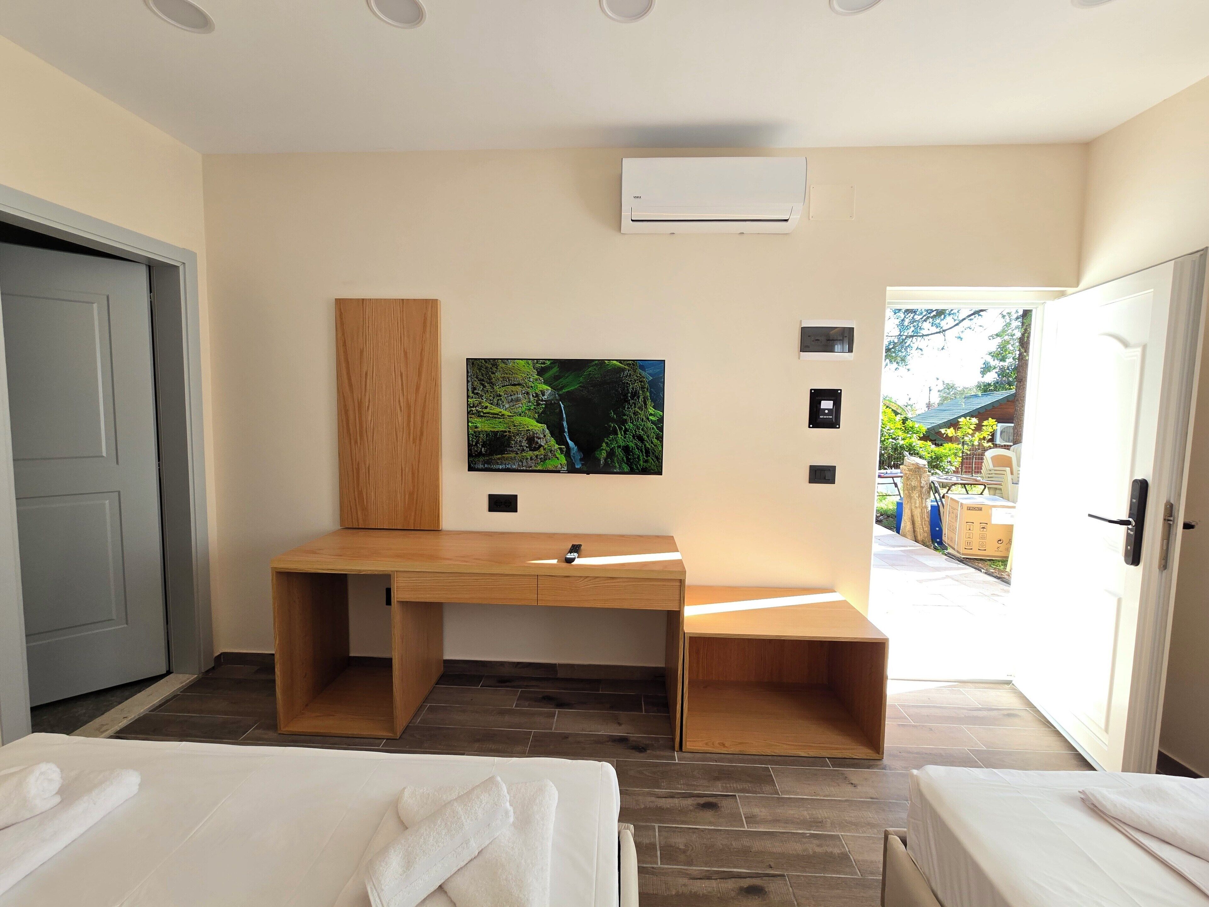 A modern hotel room with a wooden desk, a mounted TV, a small bench, and two beds.