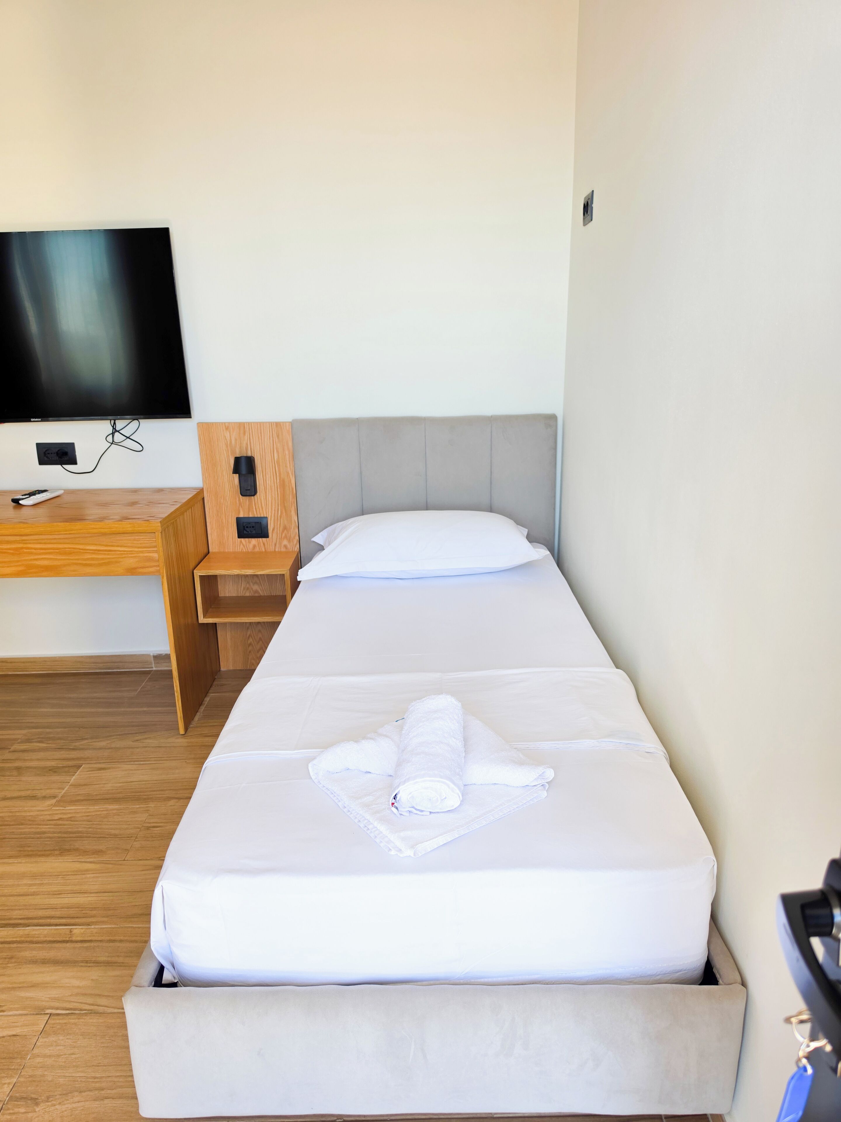 A single bed with a white mattress, a wooden nightstand, a television mounted on the wall, and a power outlet on the nightstand.