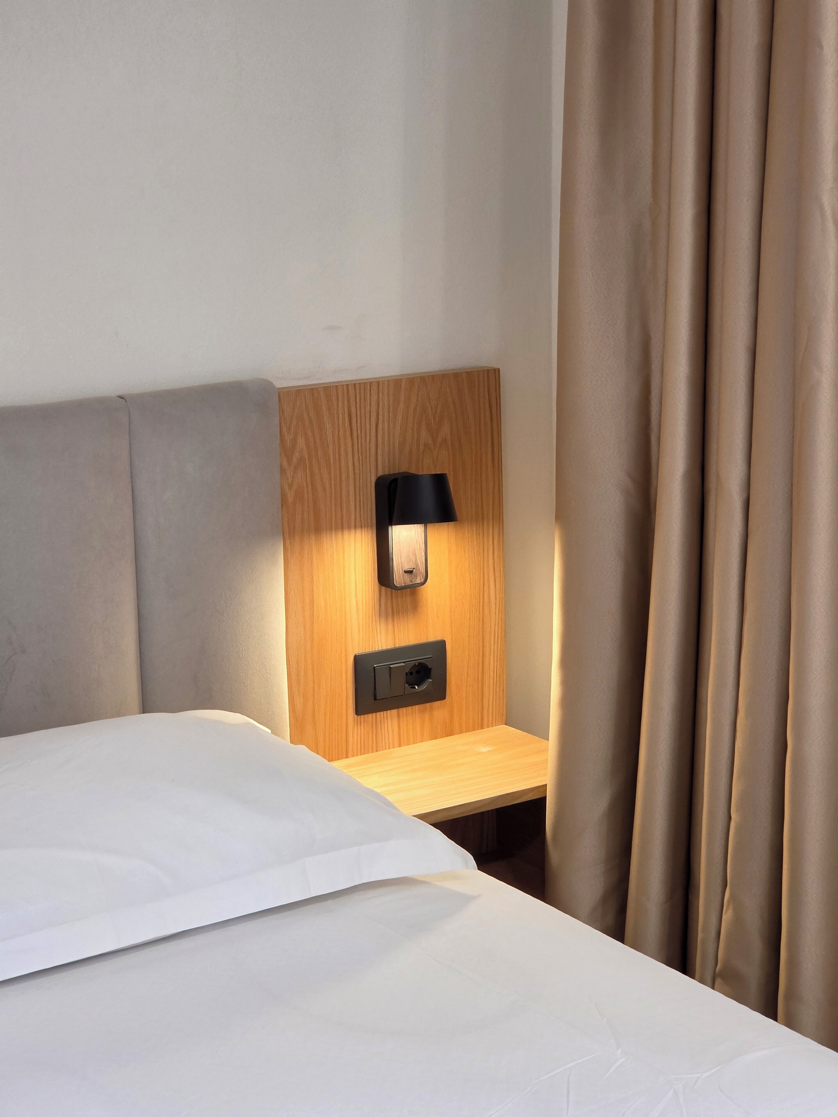 Een hotelkamer met een bed, een nachtkastje, een wandlamp en een gordijn.
